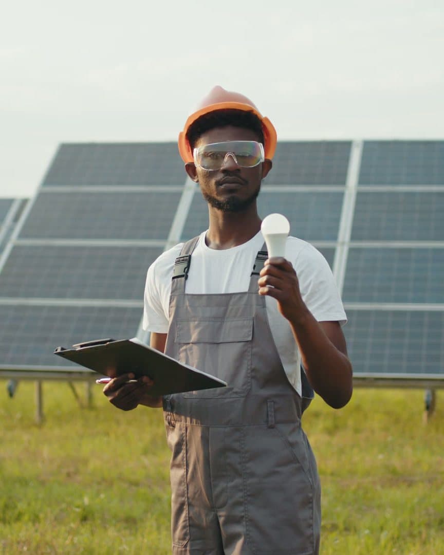 A man in overalls and an orange hard hat holds a light bulb and a clipboard in front of solar panels
