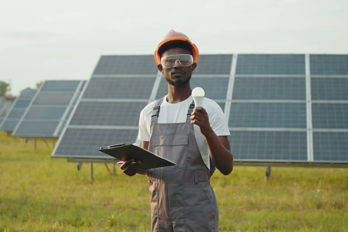 A man in overalls and an orange hard hat holds a light bulb and a clipboard in front of solar panels