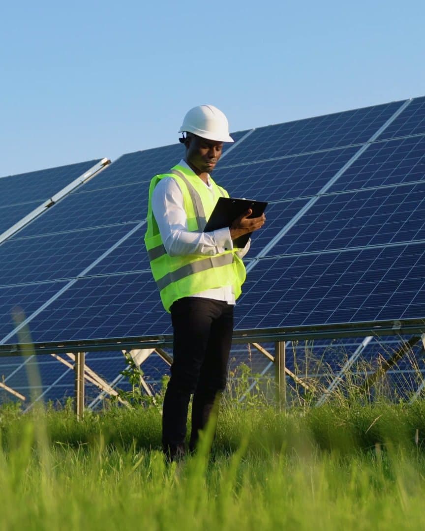 An engineer in a hard hat and high-visibility vest checks a tablet beside rows of solar panels