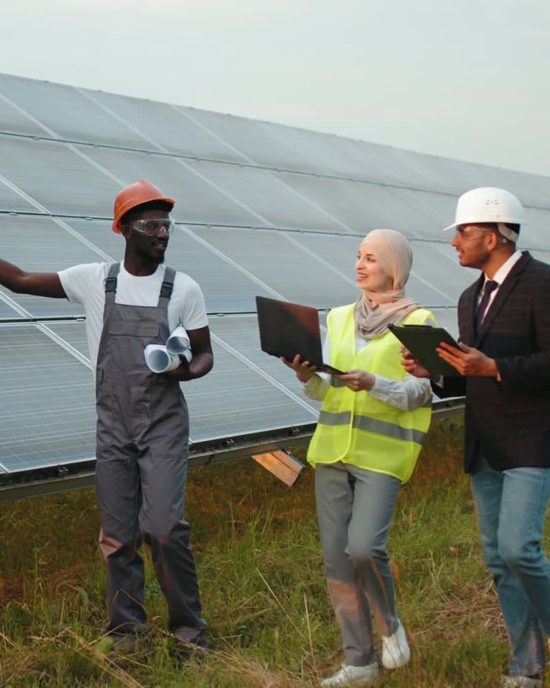 Three people in conversation beside a row of solar panels, two of them holding laptops